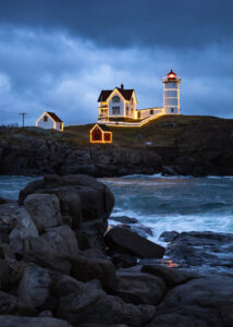 The rocky coastline sits in shadows while the holiday-decorated Nubble Lighthouse gleams against a stormy sky.